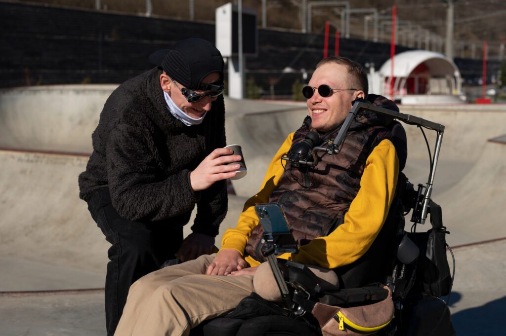 Man met ALS zit in een rolstoel in de zon en wordt geholpen door een andere man